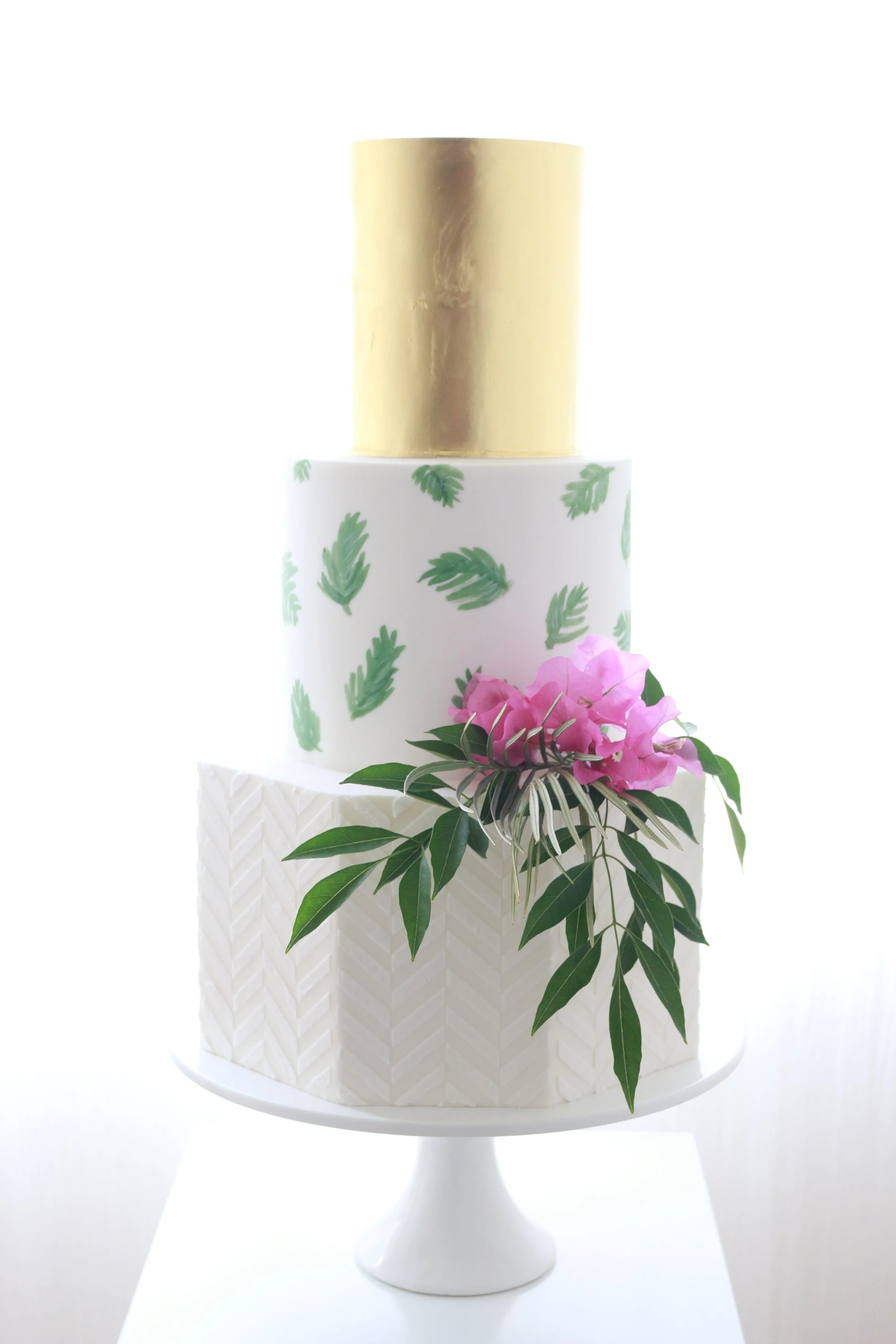 A tiered wedding cake with a white fondant base decorated with green leaves and pink flowers, topped with a smooth gold metallic finish.