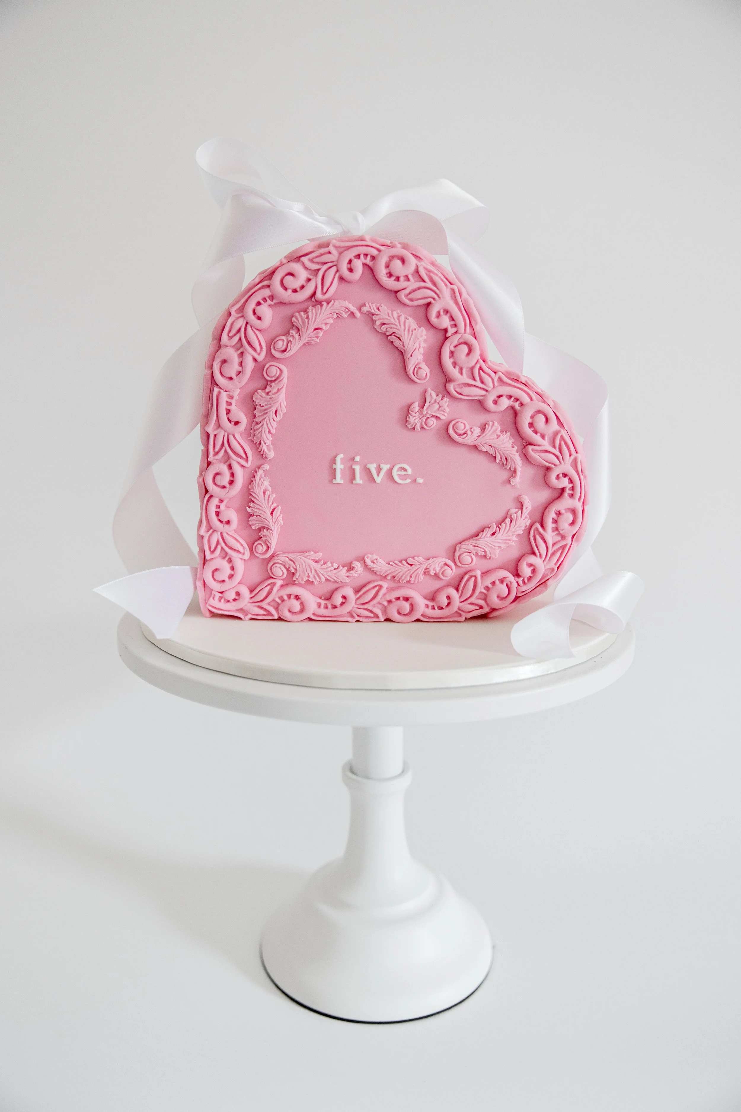 Pink embroidered oval cookie with white lettered text 'five.' and a decorative border, wrapped with a white satin ribbon, on a white cake stand.