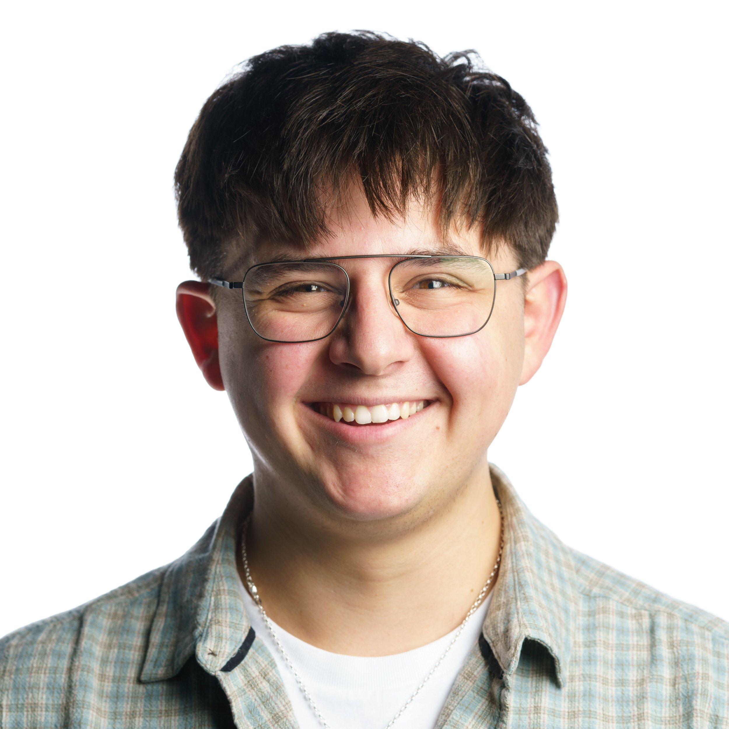 Close-up of a young man with brown curly hair, glasses, smiling, wearing a plaid shirt and a necklace.