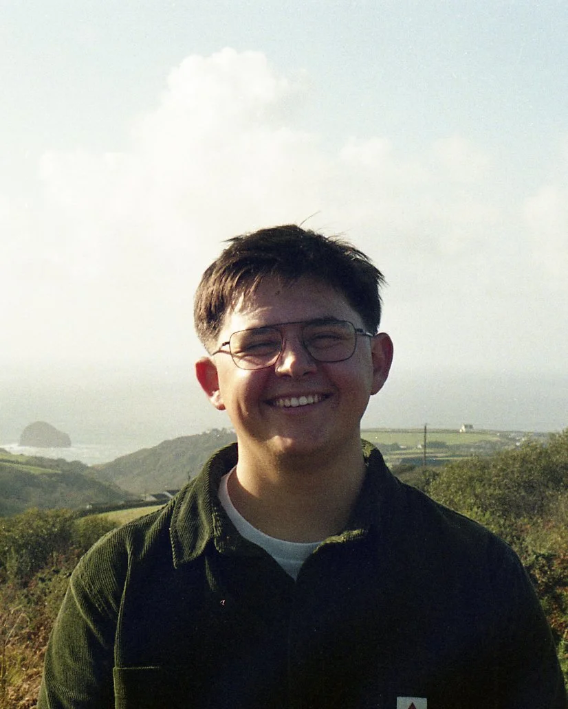 Smiling young man with glasses in an outdoor landscape with hills, cloudy sky, and distant ocean view.