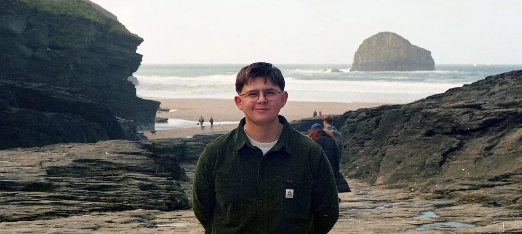 A young man with glasses and short brown hair is standing on a rocky beach with a large rock formation in the background. The weather appears cloudy or overcast.
