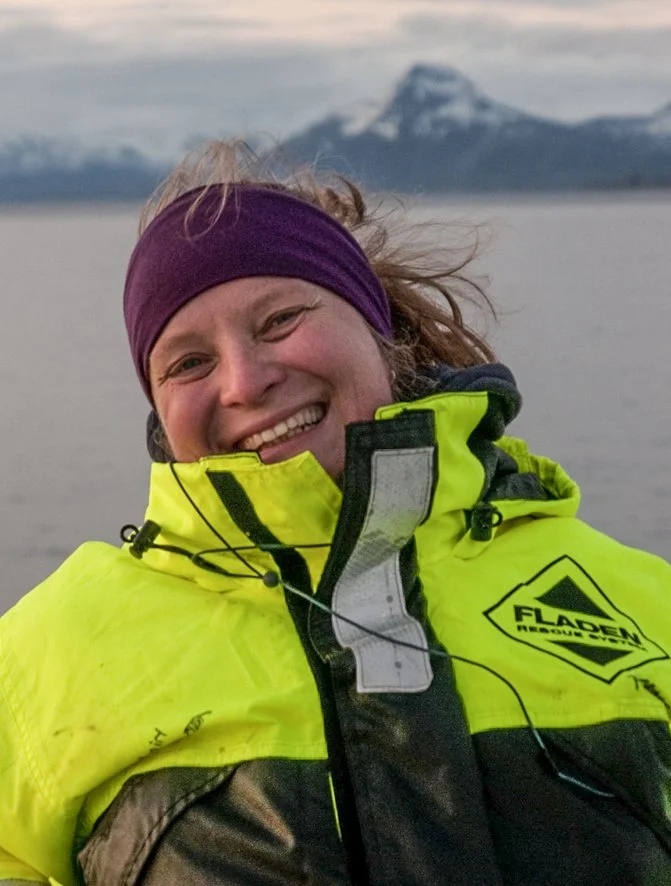 A smiling woman wearing a purple headband and a bright yellow high-visibility jacket outdoors with a mountain and body of water in the background.