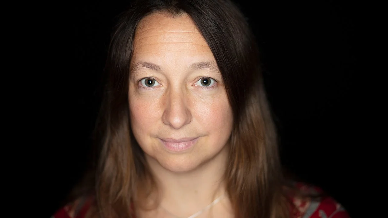 Close-up portrait of a woman with shoulder-length brown hair, blue eyes, and light skin, looking directly at the camera against a black background.