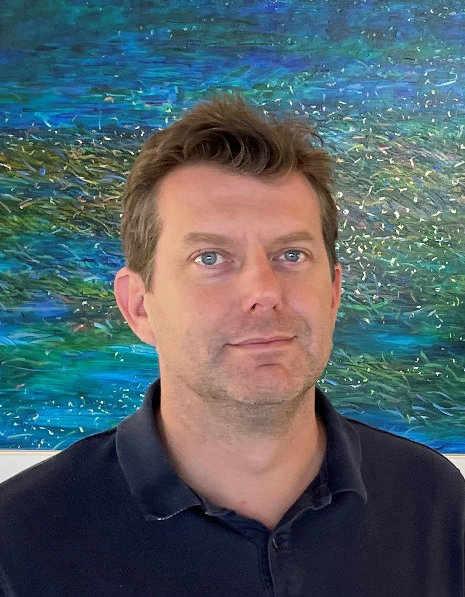 A man with short brown hair and blue eyes wearing a black collared shirt stands in front of an abstract painting with blue, green, and purple colors.