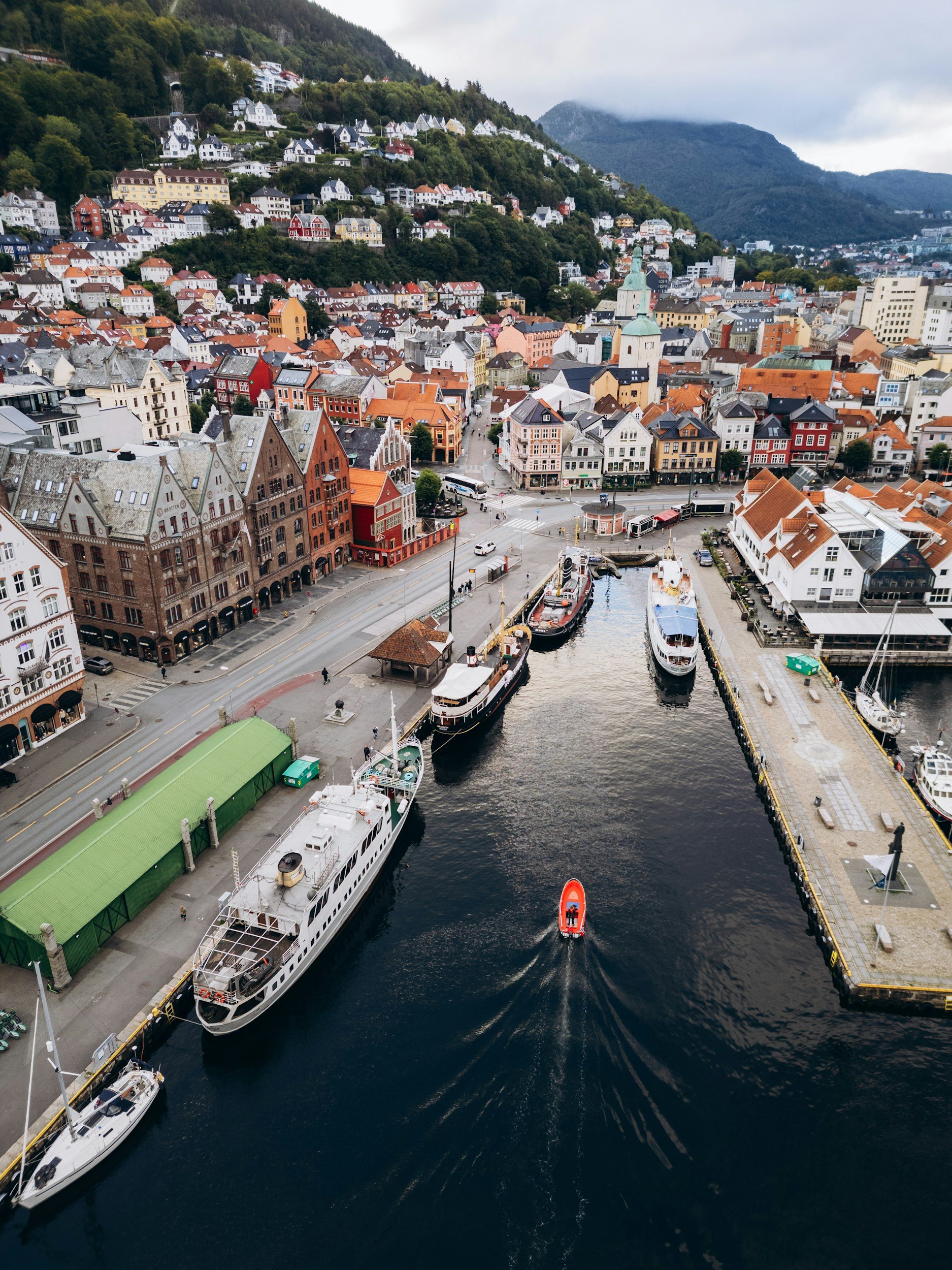 Aerial view of a harbor with boats docked, surrounded by colorful buildings, and a hilly landscape in the background.