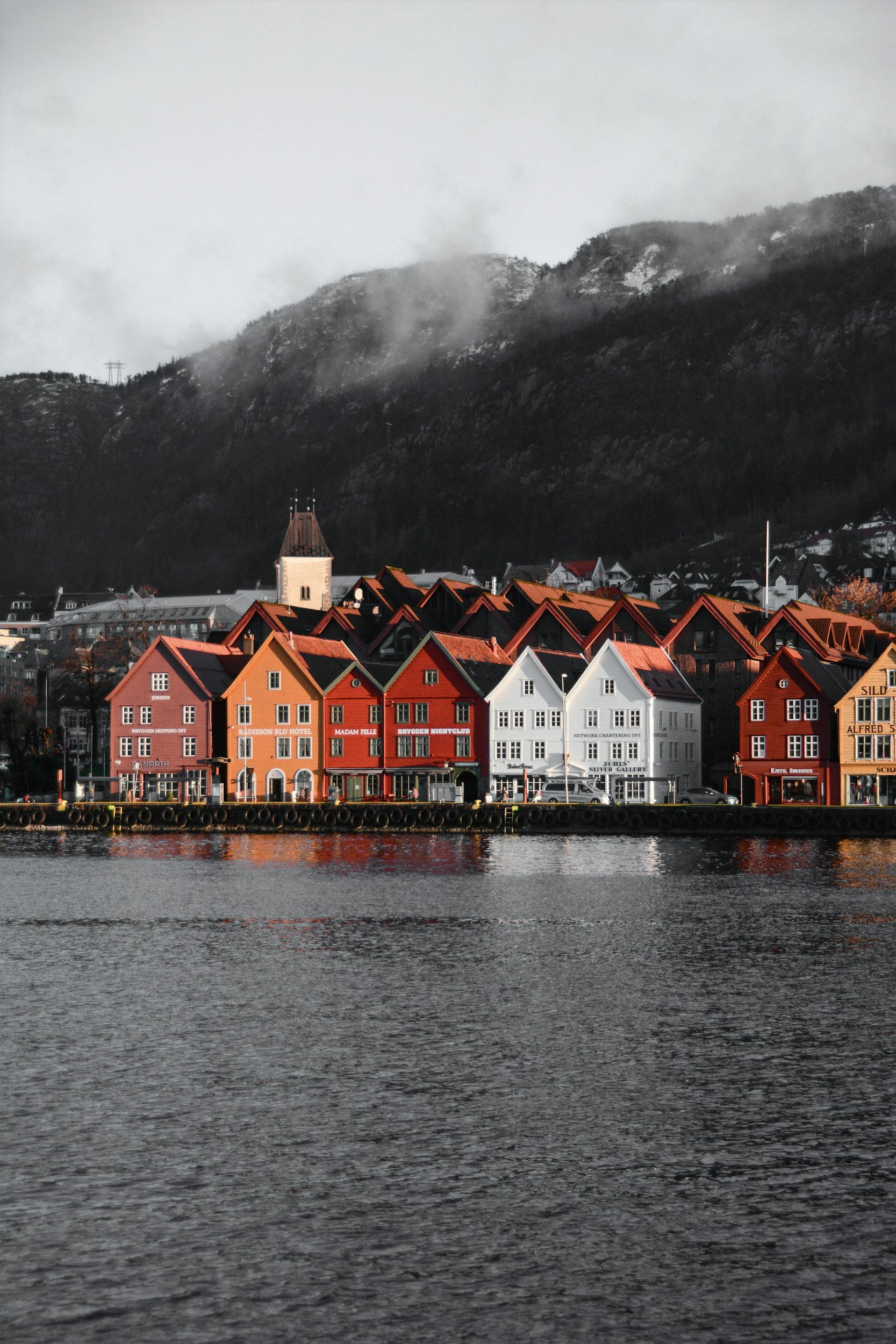 Colorful row of houses along a waterfront with mountains in the background, some clouds, and mist over the mountains.