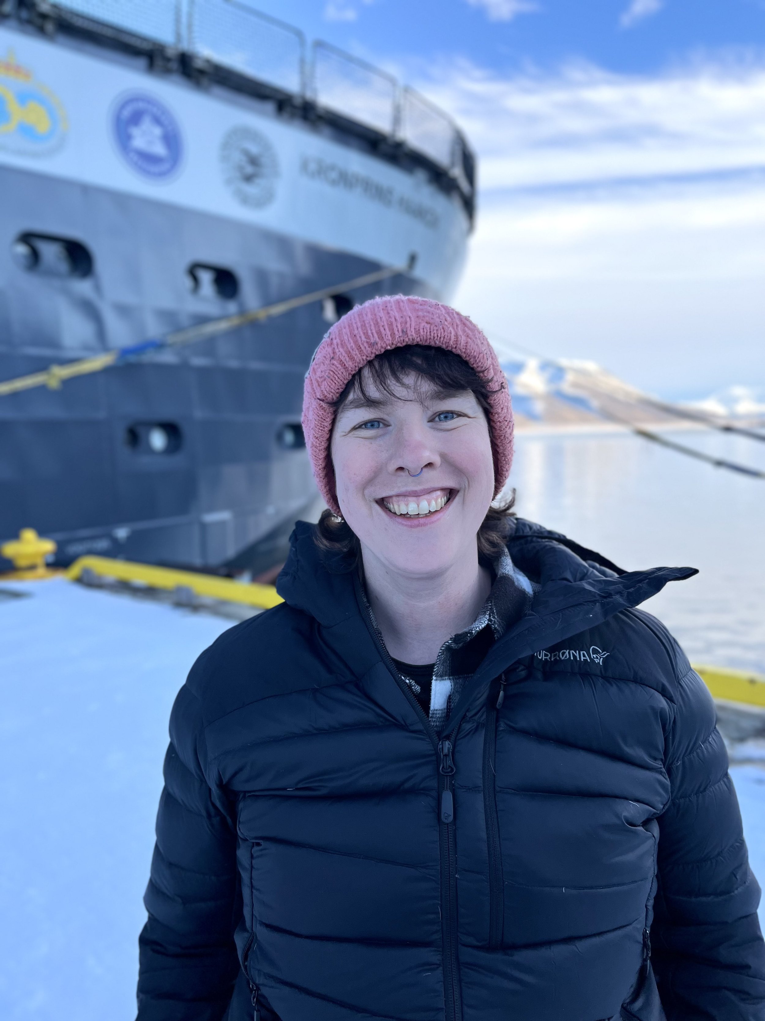 A woman smiling outdoors in front of a ship with a mountainous landscape in the background and snow on the ground.