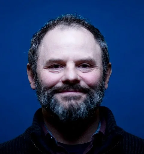 A man with short dark hair and a beard, smiling, against a dark blue background.