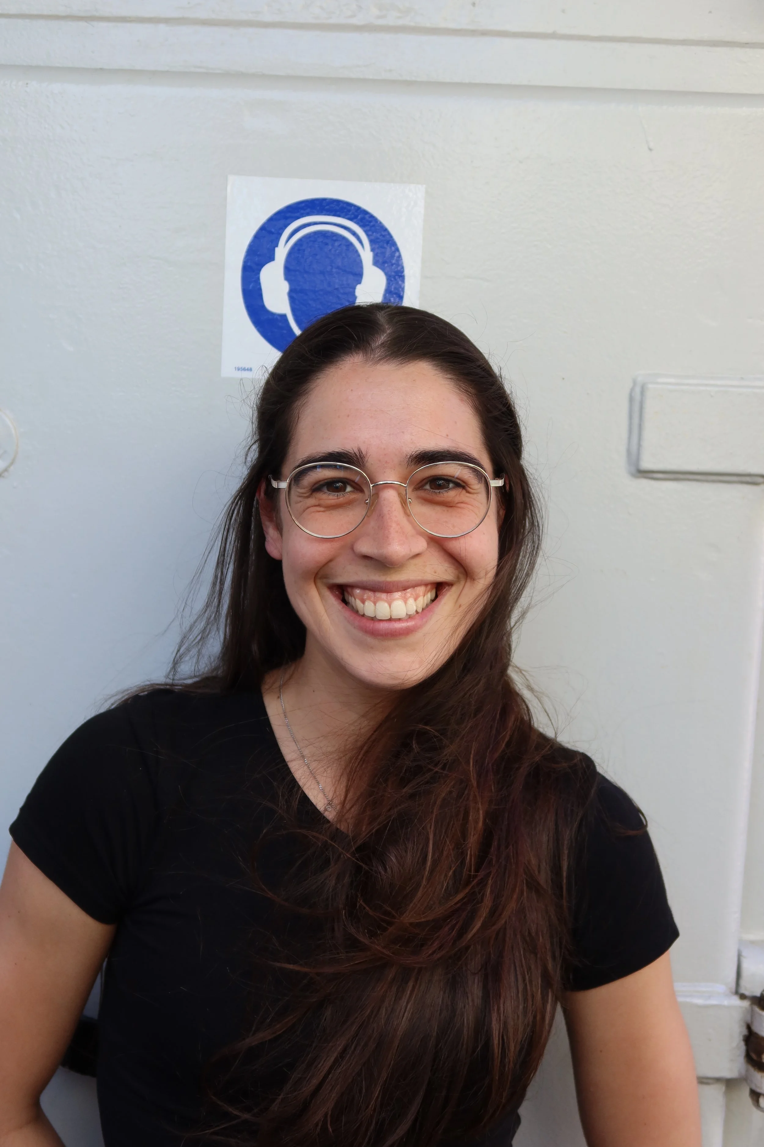 A smiling woman with long brown hair and glasses taking a selfie in front of a white wall with a blue headphone sign.