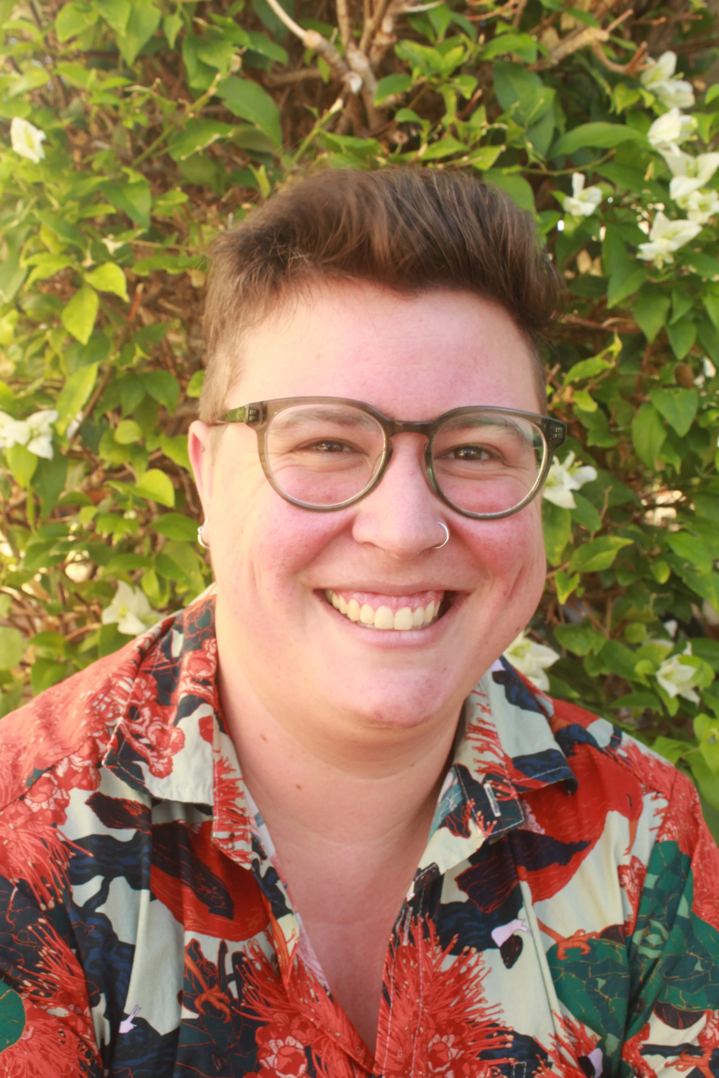 A person with short brown hair and glasses smiling outdoors in front of green leafy plants with white flowers, wearing a colorful patterned shirt.