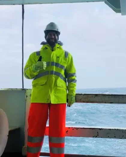 A man in a yellow safety jacket, orange pants, a white helmet, and safety gloves standing on a ship deck near the railing with the ocean in the background, giving a thumbs-up.