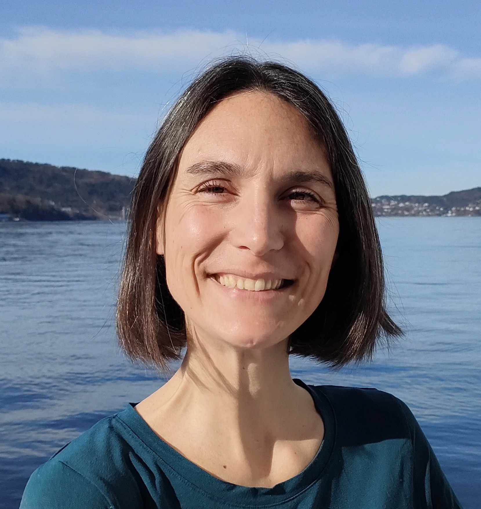 A smiling woman with straight, shoulder-length brown hair, wearing a dark top, standing outdoors near a body of water with distant hills and a partly cloudy sky in the background.