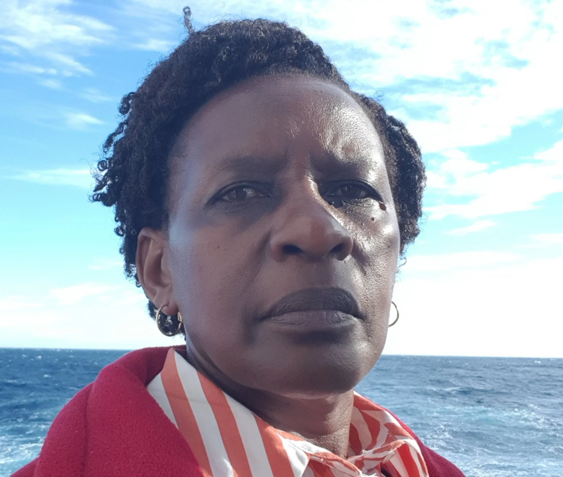 Close-up of a woman with short curly hair, wearing earrings and a red and striped shirt, standing outdoors near the water with a blue sky filled with clouds in the background.