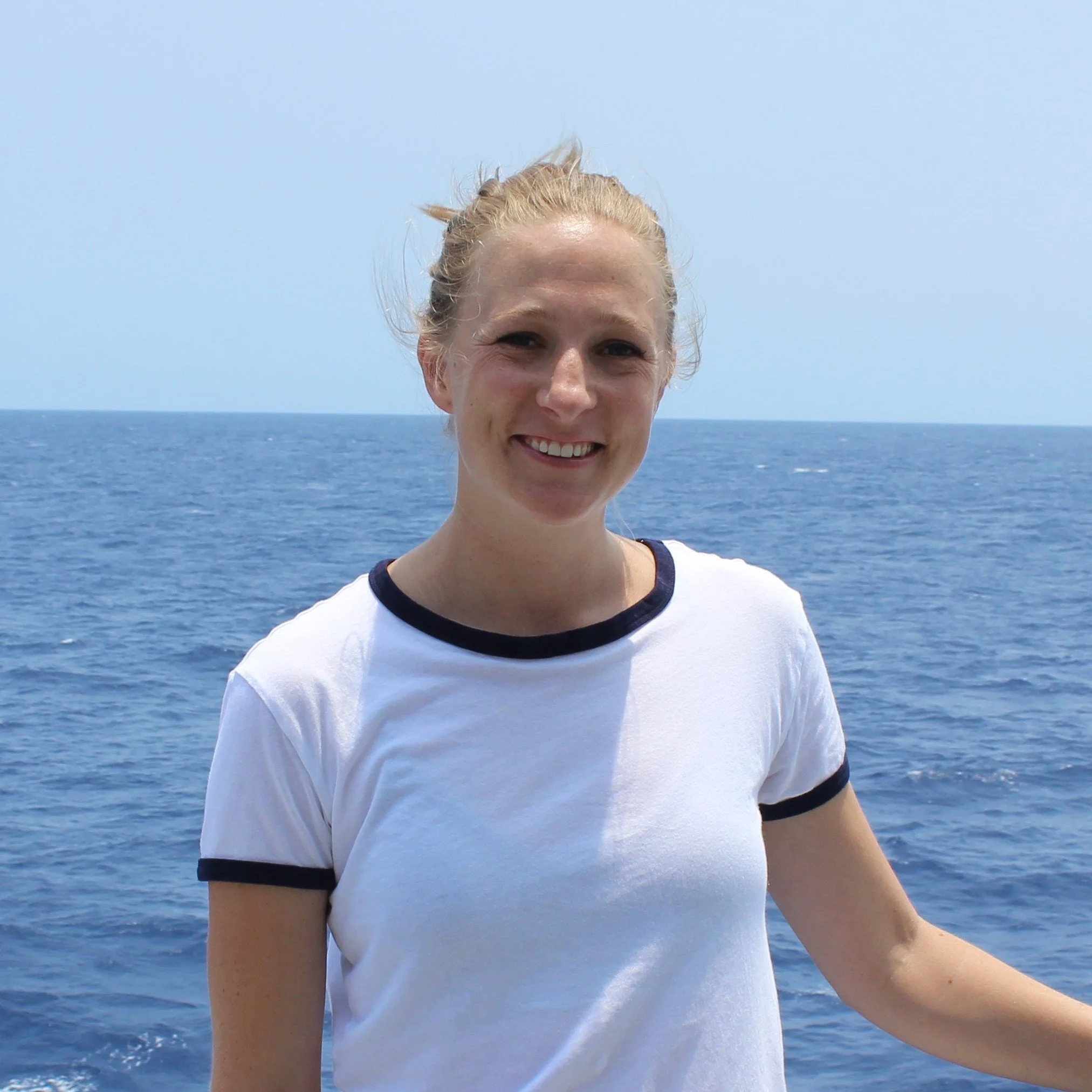 A smiling woman with blonde hair tied back, standing over the ocean with a clear blue sky.