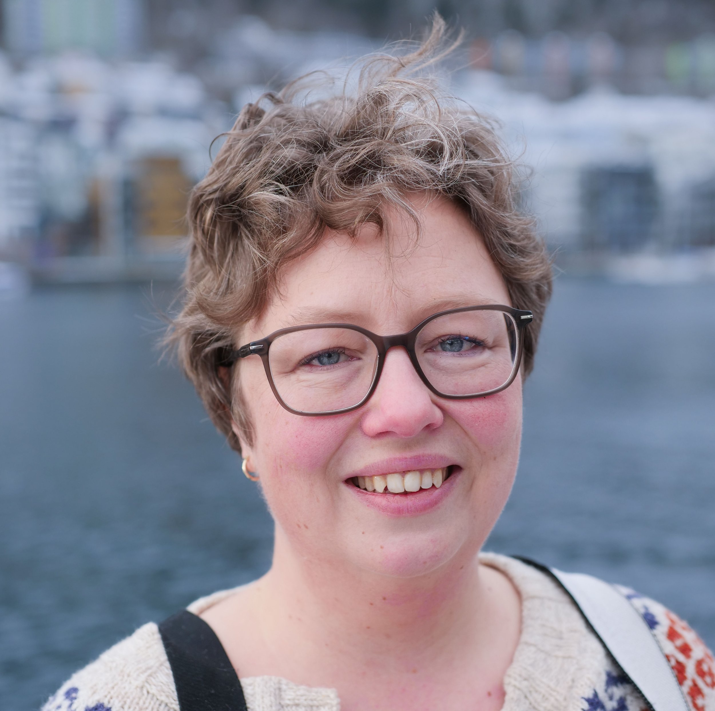 Close-up of a person's face, focusing on their eyes, glasses, hair, and smile. The person has short, curly hair and light-colored eyes, with a blurred background.