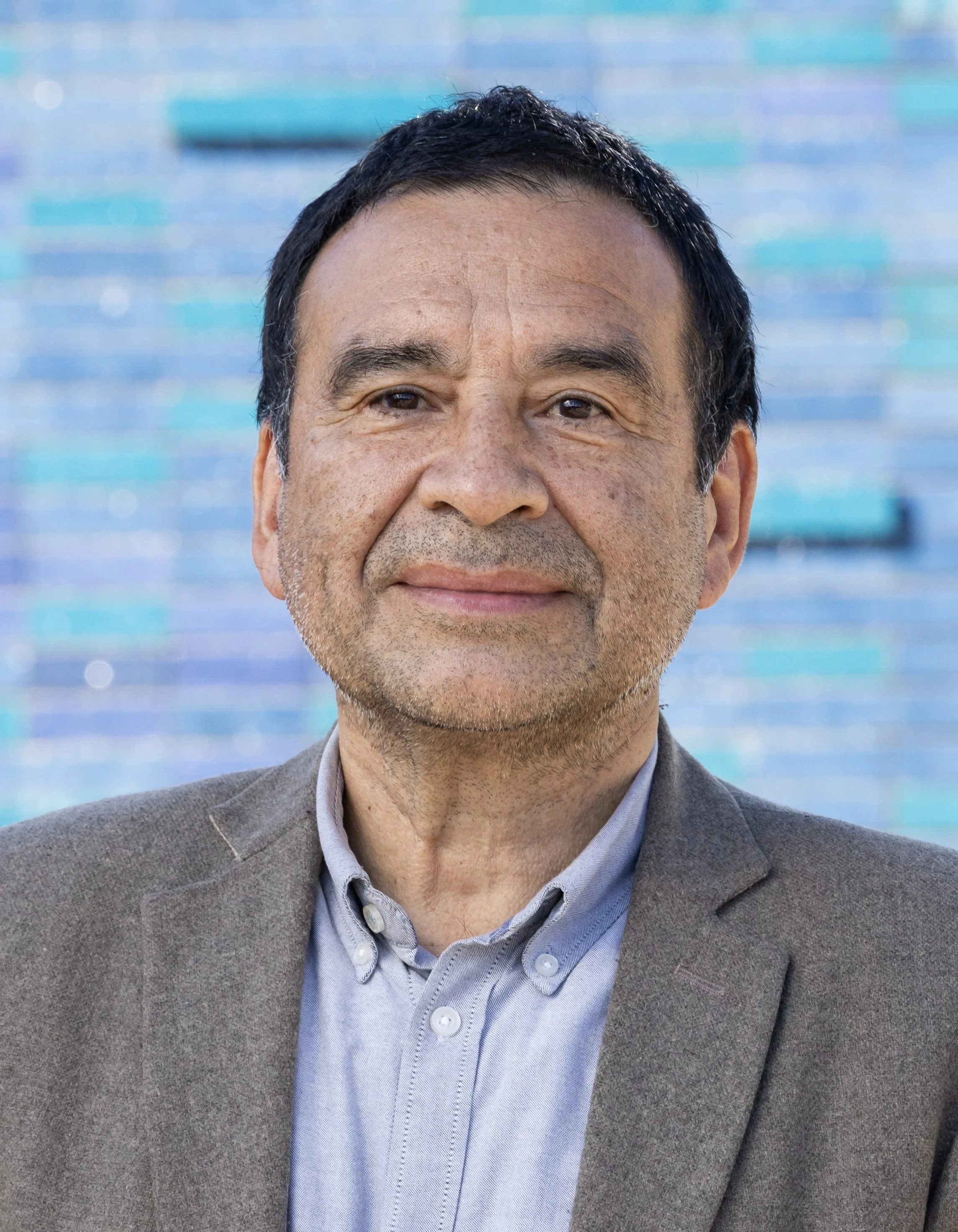 Close-up of a man with dark hair, smiling, wearing a gray blazer and light blue shirt against a blue and green blurred background.