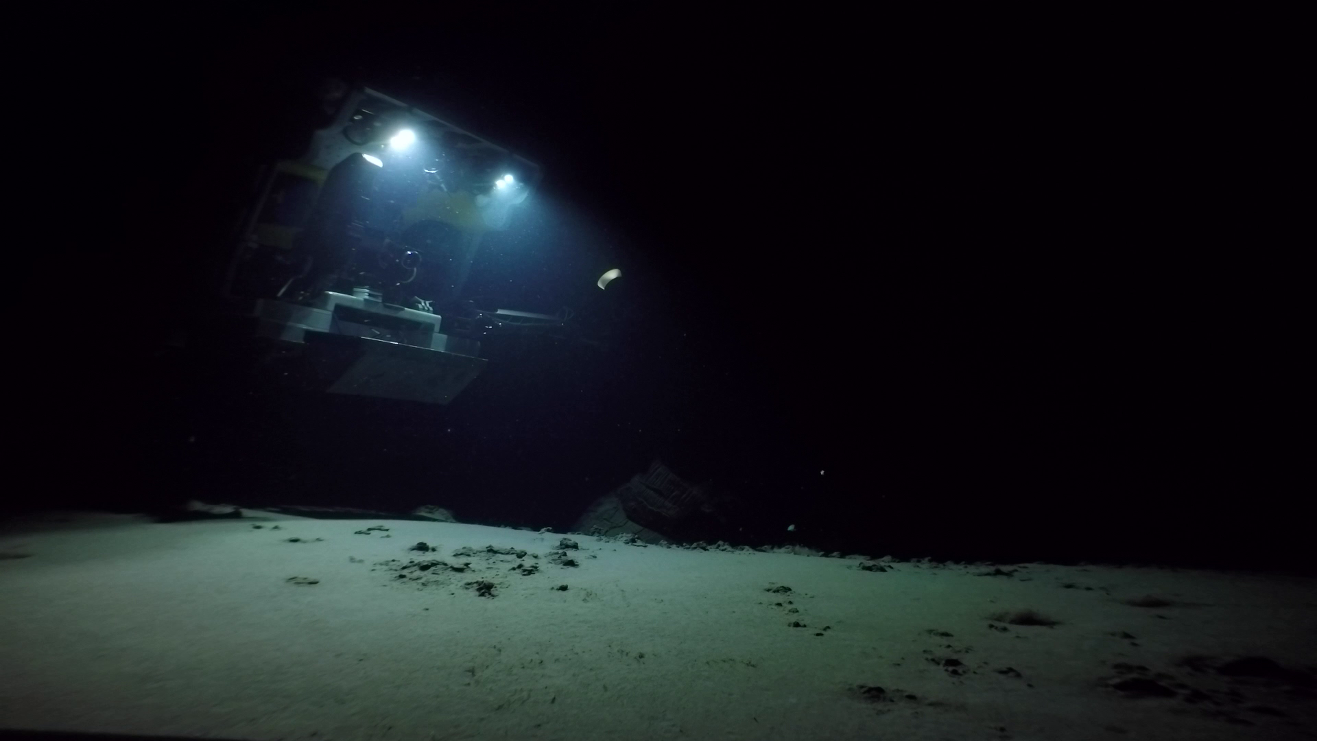 Underwater scene with an ROVresting on the ocean floor, illuminated by lights, with visible sandy seabed and rocks.
