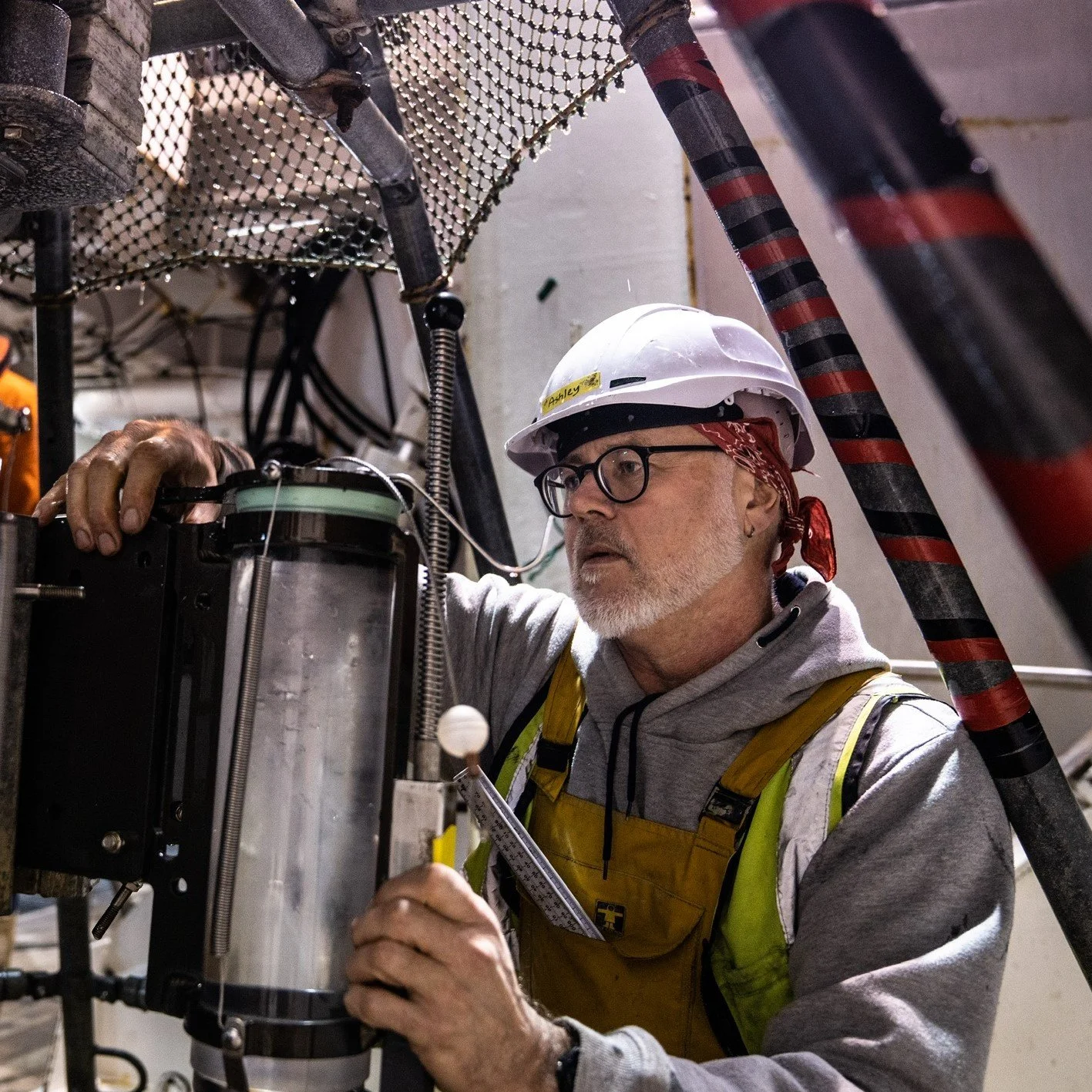 A man with a safety helmet, glasses, and a yellow safety vest working on equipment indoors.