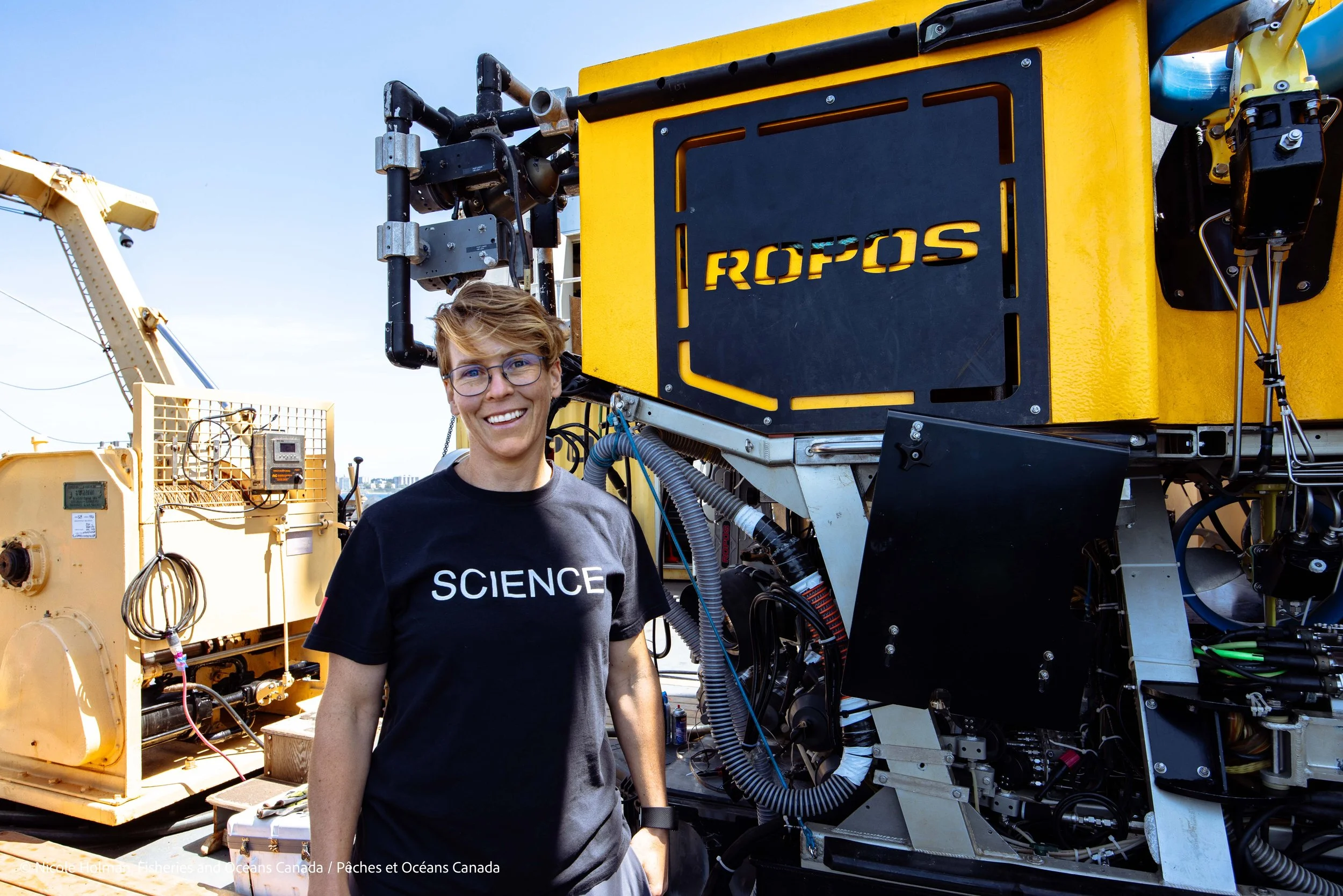 A woman with short hair and glasses smiling stands next to scientific or technical equipment labeled 'ROPOS'. She wears a black T-shirt with the word 'SCIENCE' on it, and is outside under a clear sky.
