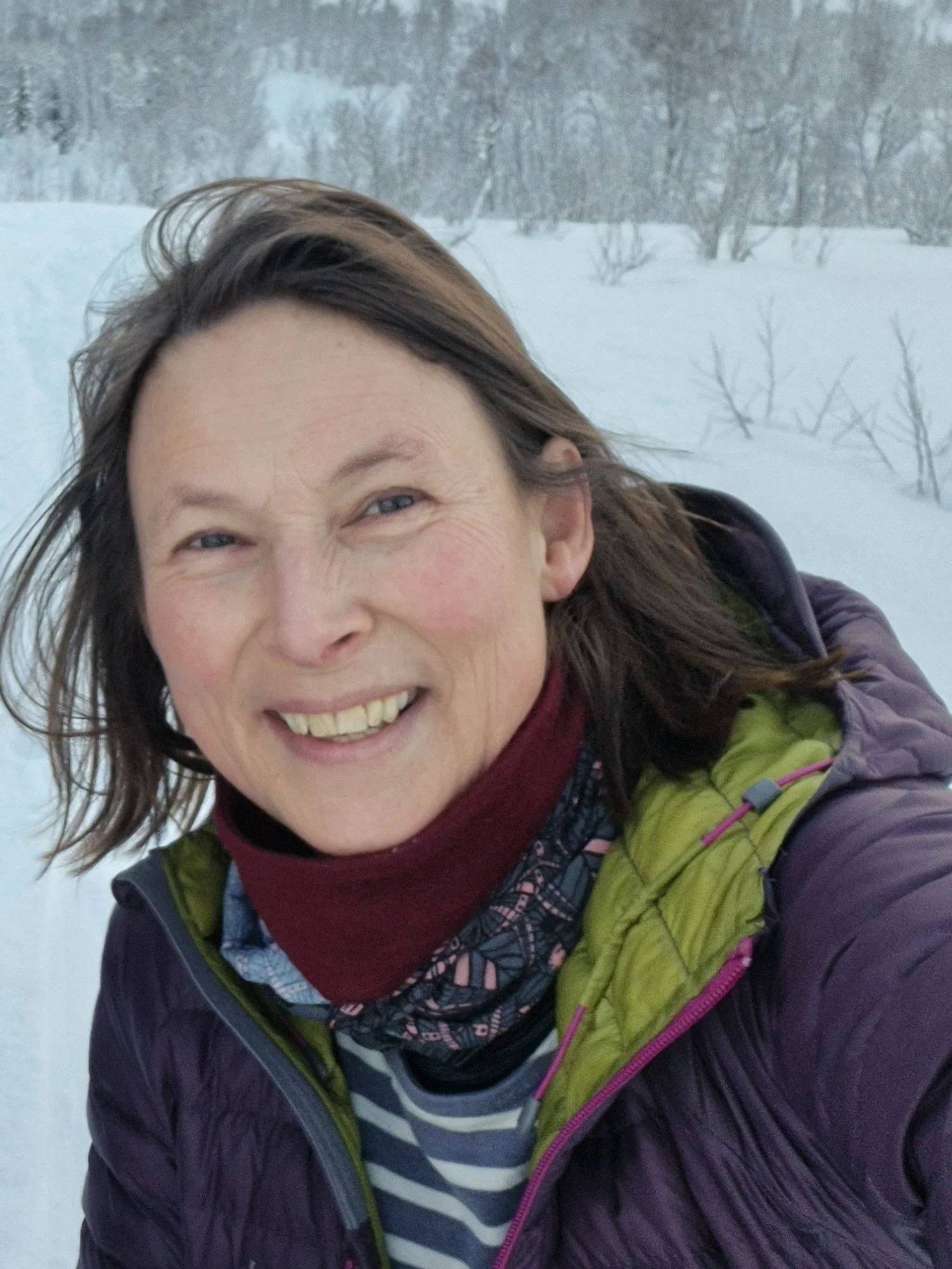 A woman smiling outdoors in a snowy landscape, wearing a purple jacket, a green vest, a patterned scarf, and a red neck warmer.