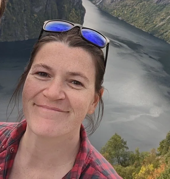 A woman taking a selfie outdoors overlooking a river canyon with steep cliffs and green trees.