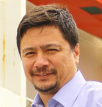 A man with dark hair, a beard, and a mustache, wearing a light blue shirt, smiling outdoors.