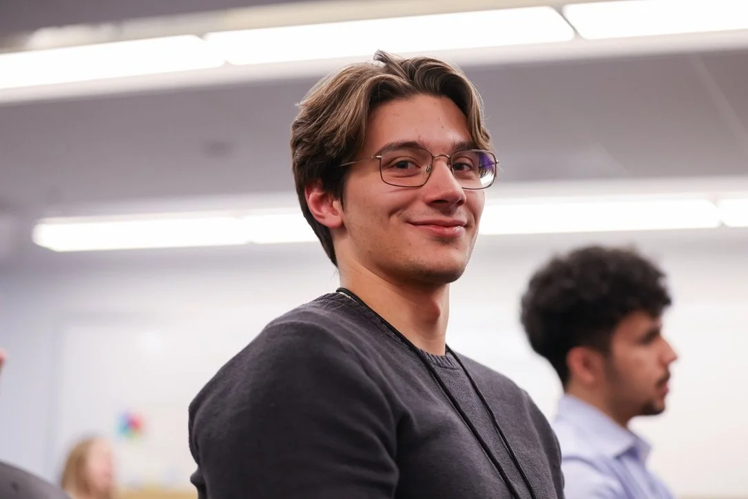 Igor Serafini. A young man with brown hair, glasses, and a black shirt, smiling in a conference room, with other people in the background.