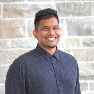 Dr. Gerome Manson. A smiling man with short dark hair wearing a dark button-up shirt against a textured gray stone wall.