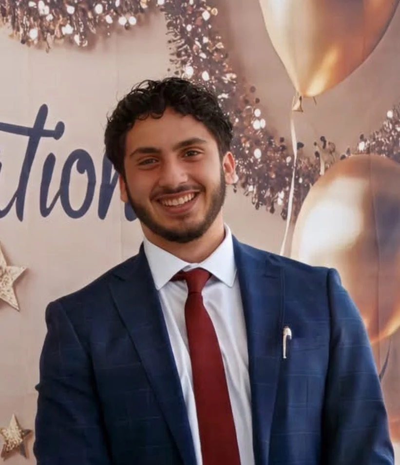 Abed A. Hijleh. A young man with dark, curly hair smiling in front of festive decorations, including gold balloons and star-shaped ornaments, with a background that has the partial word 'Graduation' and glittery decorations.