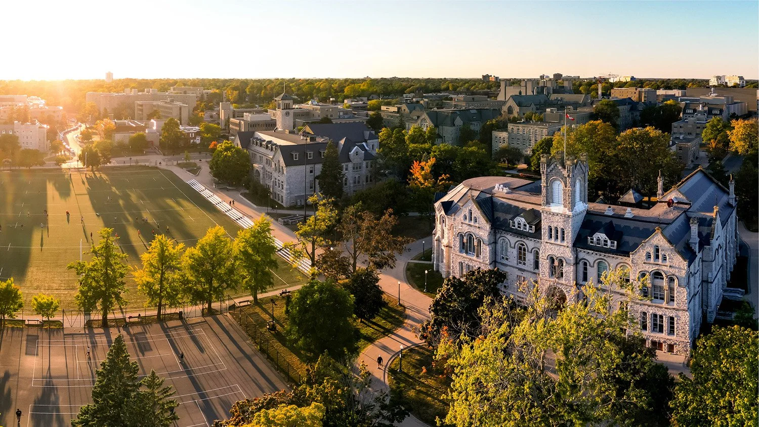 Aerial view of a historic building on a Queen's University campus with trees, Nixon field, and a city skyline in the background during sunset.