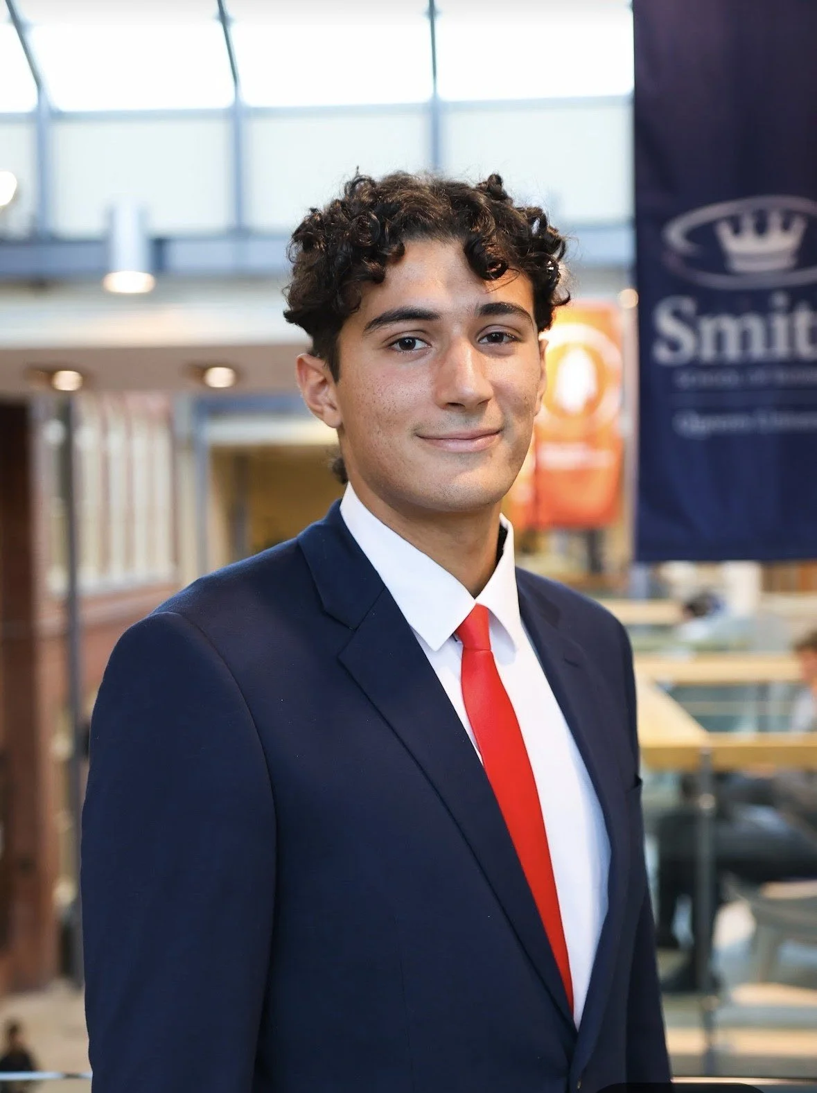 Andrei Neder. Young man in formal business attire, wearing a navy suit and red tie, smiling in a modern indoor setting with a Smith banner in the background.
