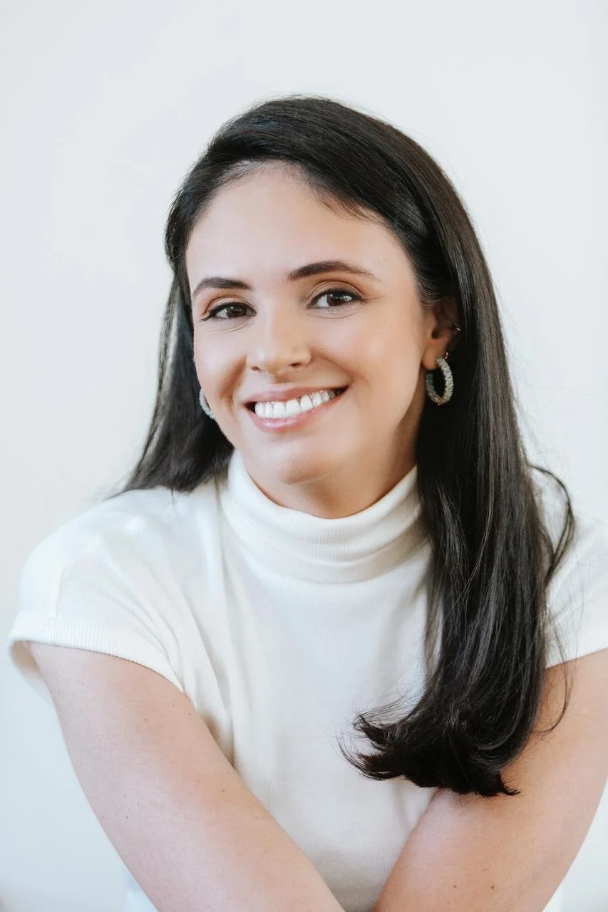 Mulher com cabelos longos e escuros, sorrindo, usando brinco de argola e blusa branca de gola alta, fundo branco.
