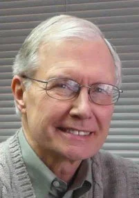 An older man with glasses and gray hair smiling in front of a window with blinds.