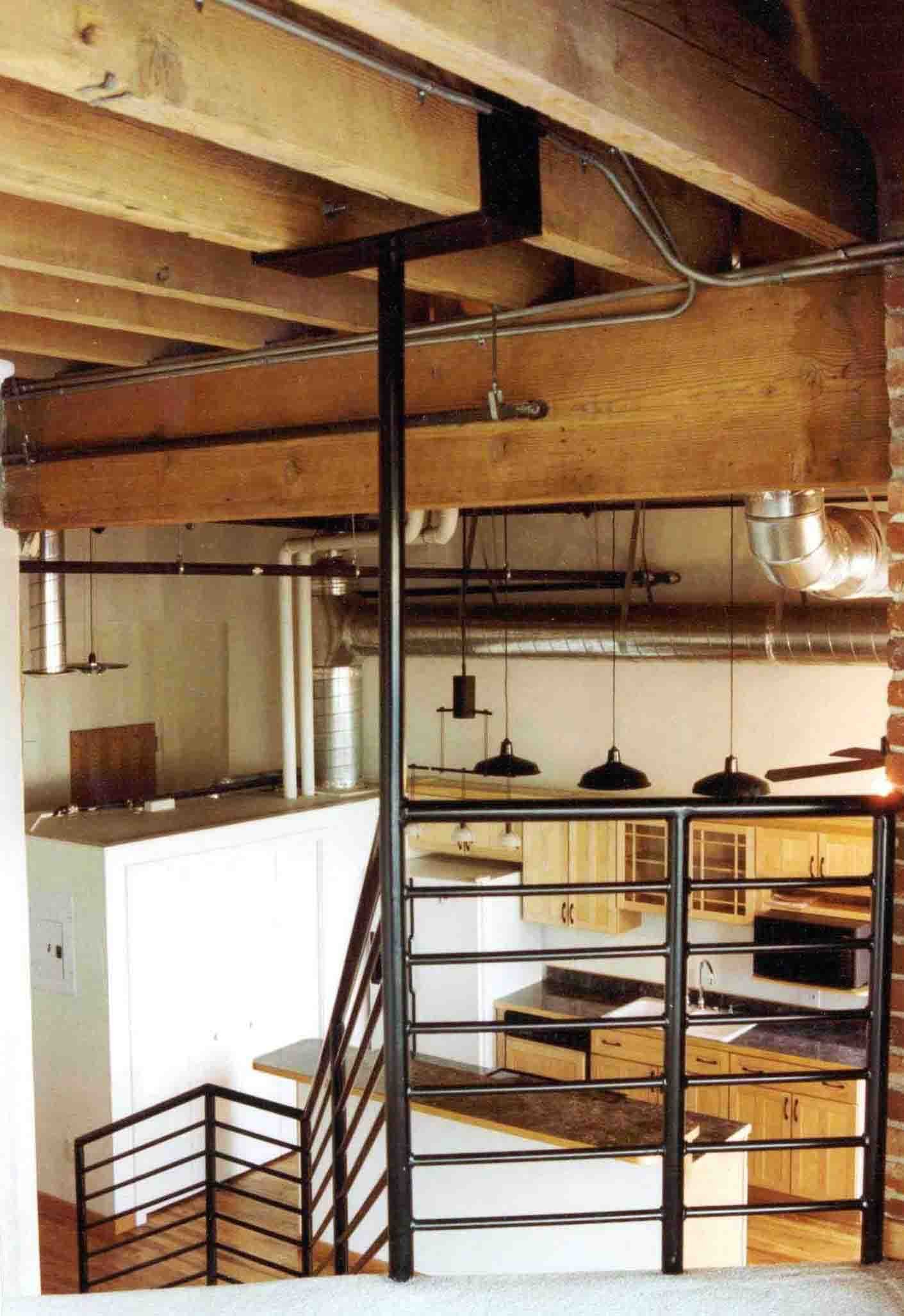 View of a staircase with black metal railing leading down to a lower level in a home with an open kitchen and living area. The kitchen has wooden cabinets, a white countertop, and metal ductwork on the ceiling.