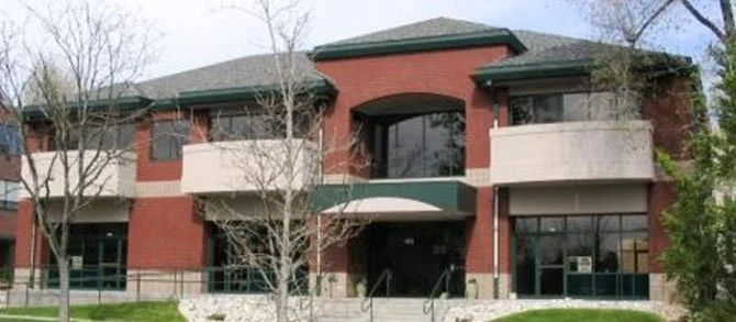 A two-story brick building with large windows and balconies, surrounded by trees and a sidewalk.