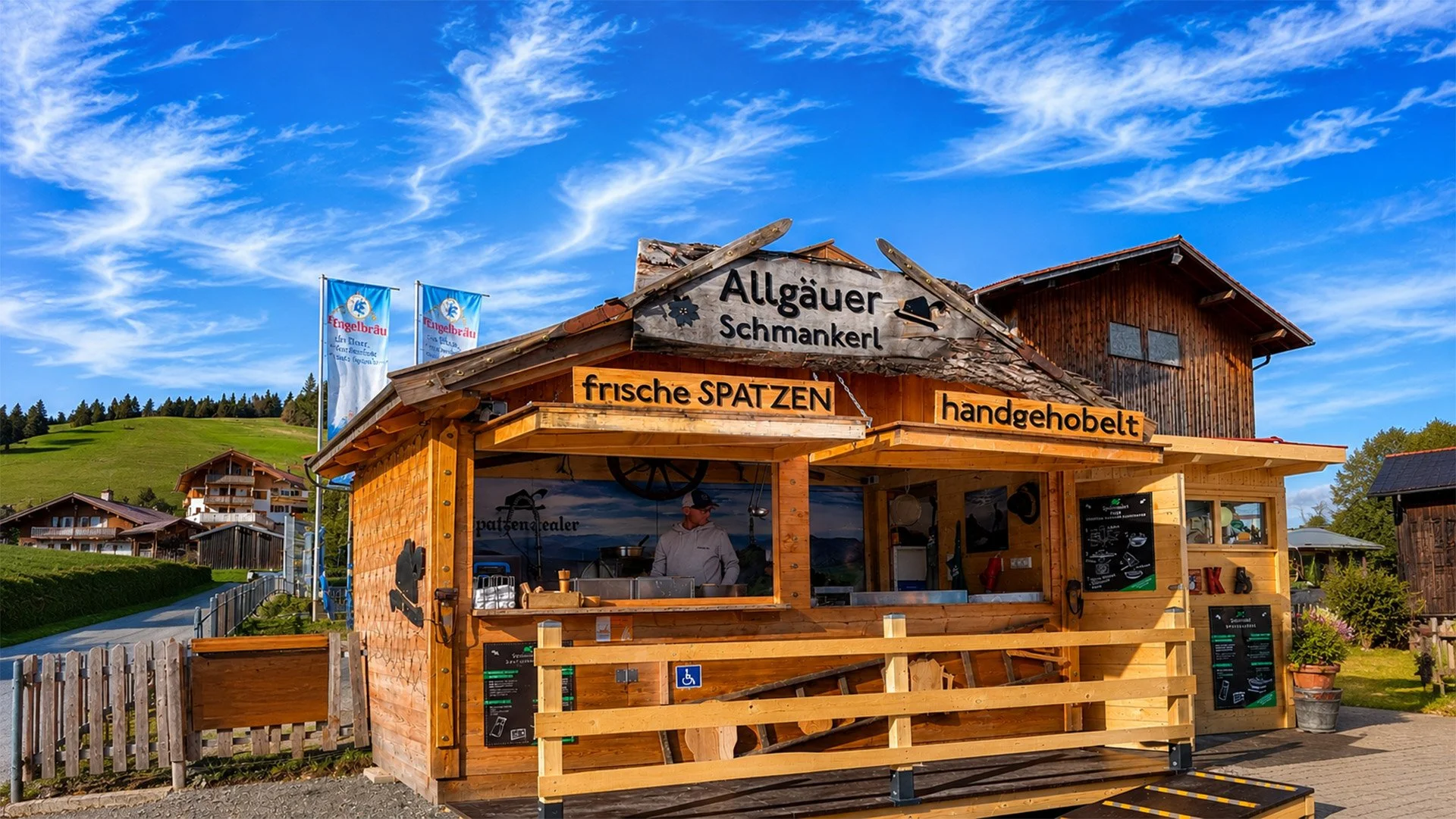 Ein Holzverkaufsstand mit der Beschriftung "Allgäuer Schmankerl", der frische Spatzen und handgehobelte Produkte anbietet, mit einem Mann, der darin arbeitet, und einem ländlichen Hintergrund mit grünen Hügeln und Häusern.