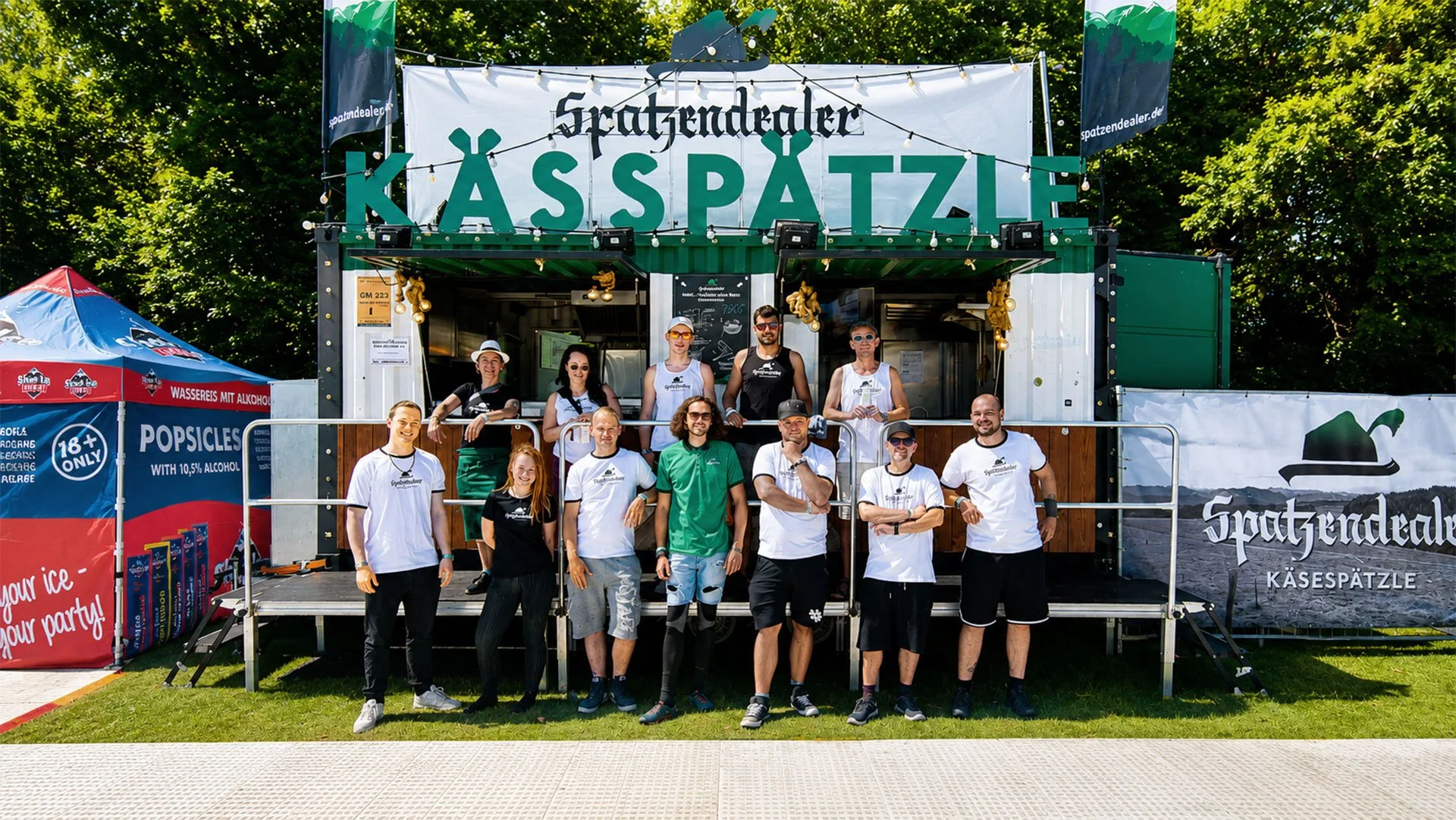 Gruppe von Menschen vor einem Verkaufsstand für Käsespätzle bei einem Festival oder Event im Freien, mit grünen Bäumen im Hintergrund.