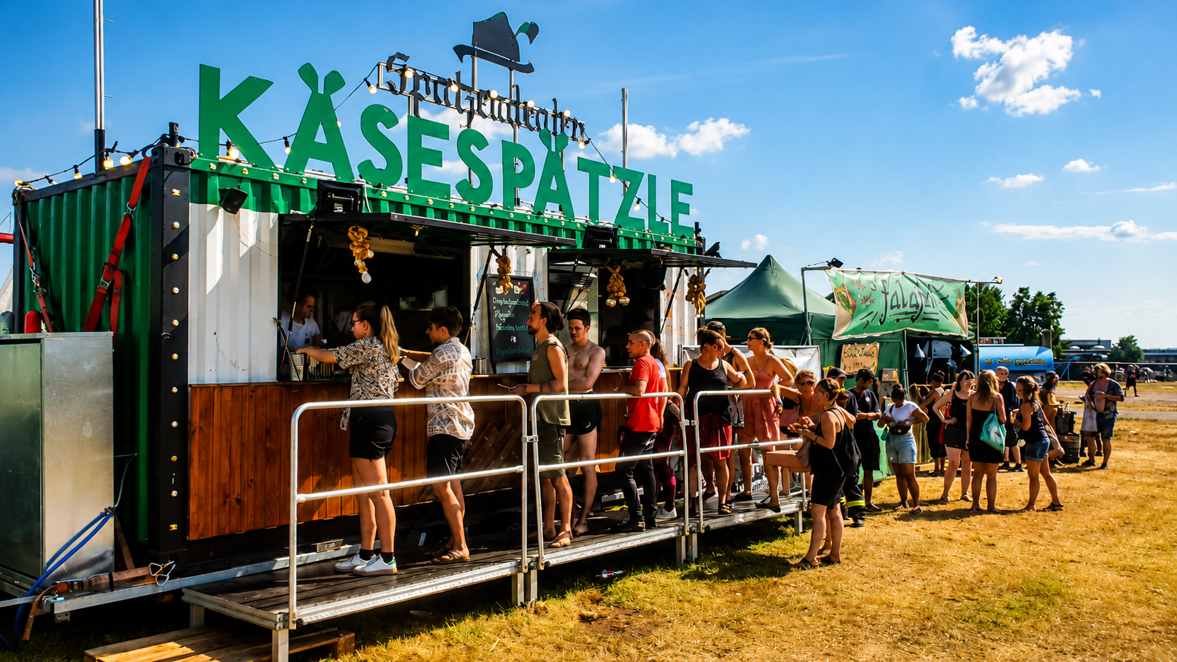 Menschen stehen in einer Linie vor einem Kassenstand mit dem Schild 'Kassenspätzle' auf dem Festival. Es gibt Tische und Stände im Freien bei sonnigem Wetter.