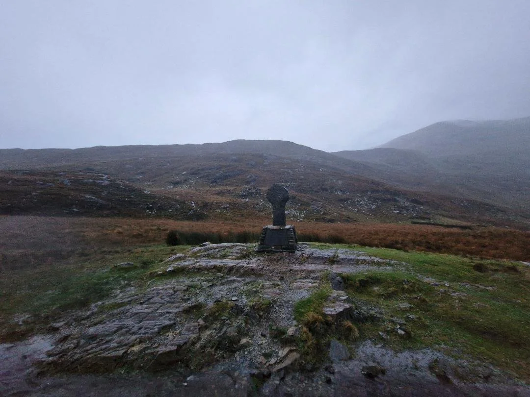 A dark stone sculpture on a pedestal in a misty outdoor landscape with rolling hills and grassy terrain.