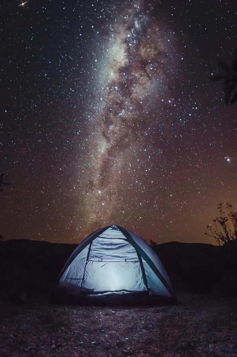 Two people sitting around a campfire under a starry night sky with the Milky Way galaxy visible.