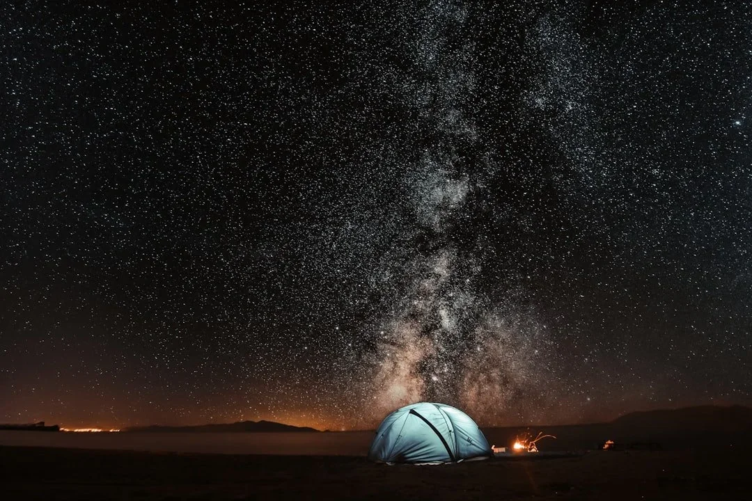 A night sky filled with countless stars and the Milky Way galaxy visible above a camping tent on a mountain landscape.