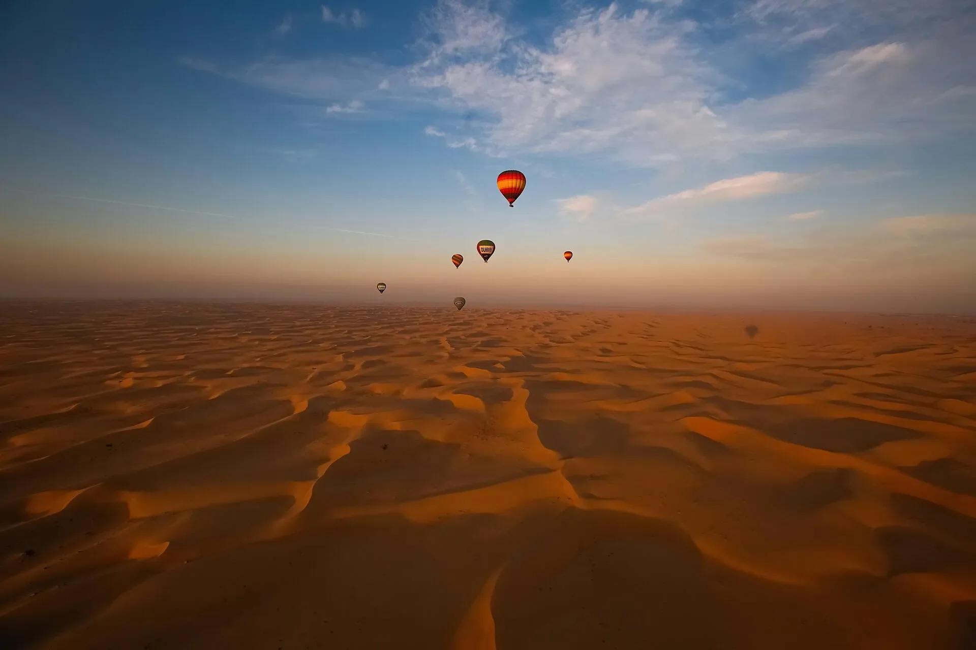 Multiple hot air balloons floating over a desert landscape during sunset with orange sand dunes and a partly cloudy sky.