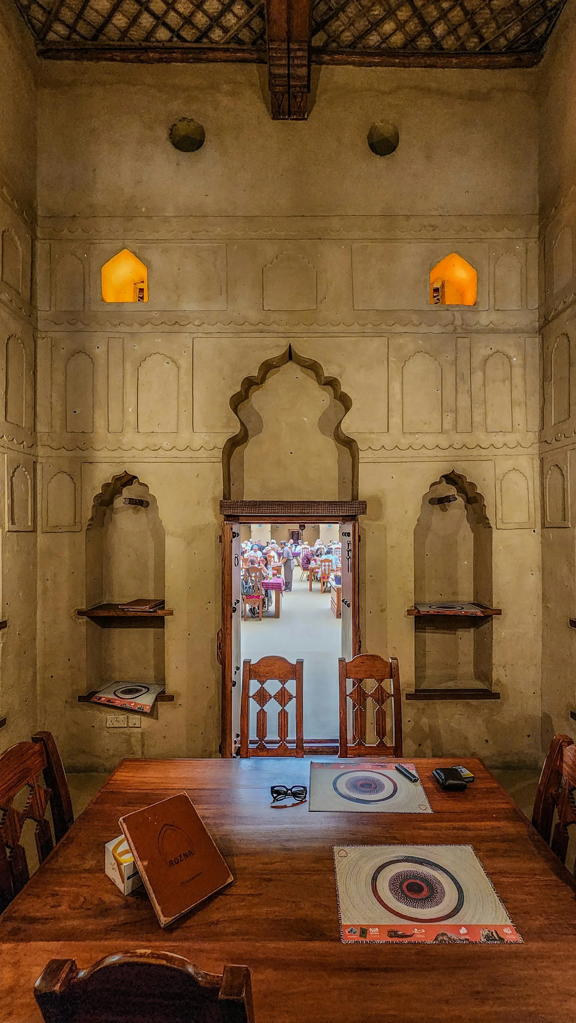 The image depicts a room with beige, textured walls and wooden furniture. There is a doorway in the wall, leading to a busy kitchen with people preparing food. The wall features architectural details like arches and decorative lines. On the floor and furniture are various magazines, pamphlets, and electronic devices, suggesting a casual or waiting area.