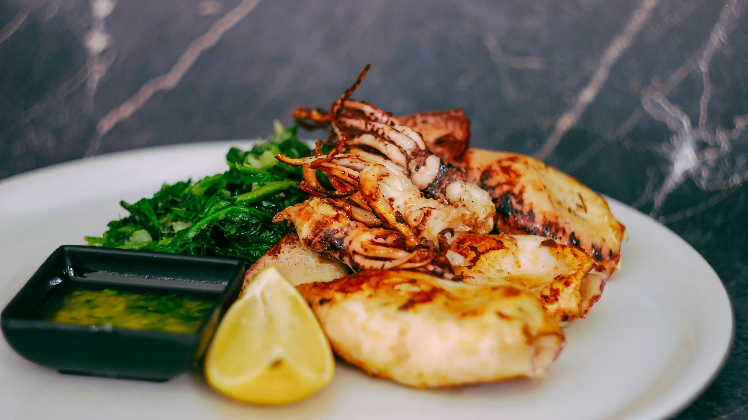 Grilled fish fillet served with grilled squid, a side of cooked greens, a lemon wedge, and a small dish of green sauce.