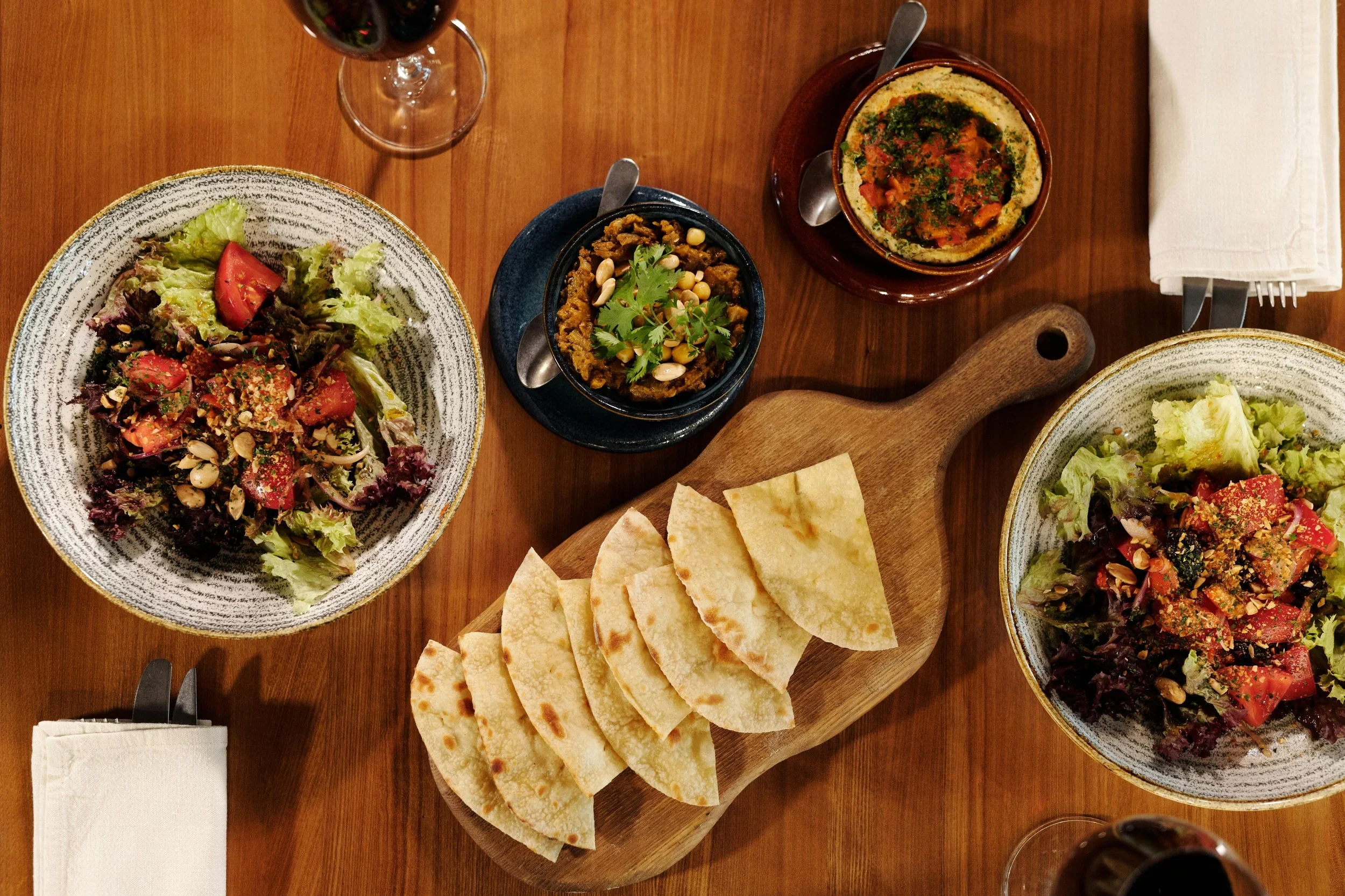 A top-down view of a meal with two salads, a bowl of lentil stew, a bowl of hummus, a stack of pita bread, and a glass of red wine on a wooden table.