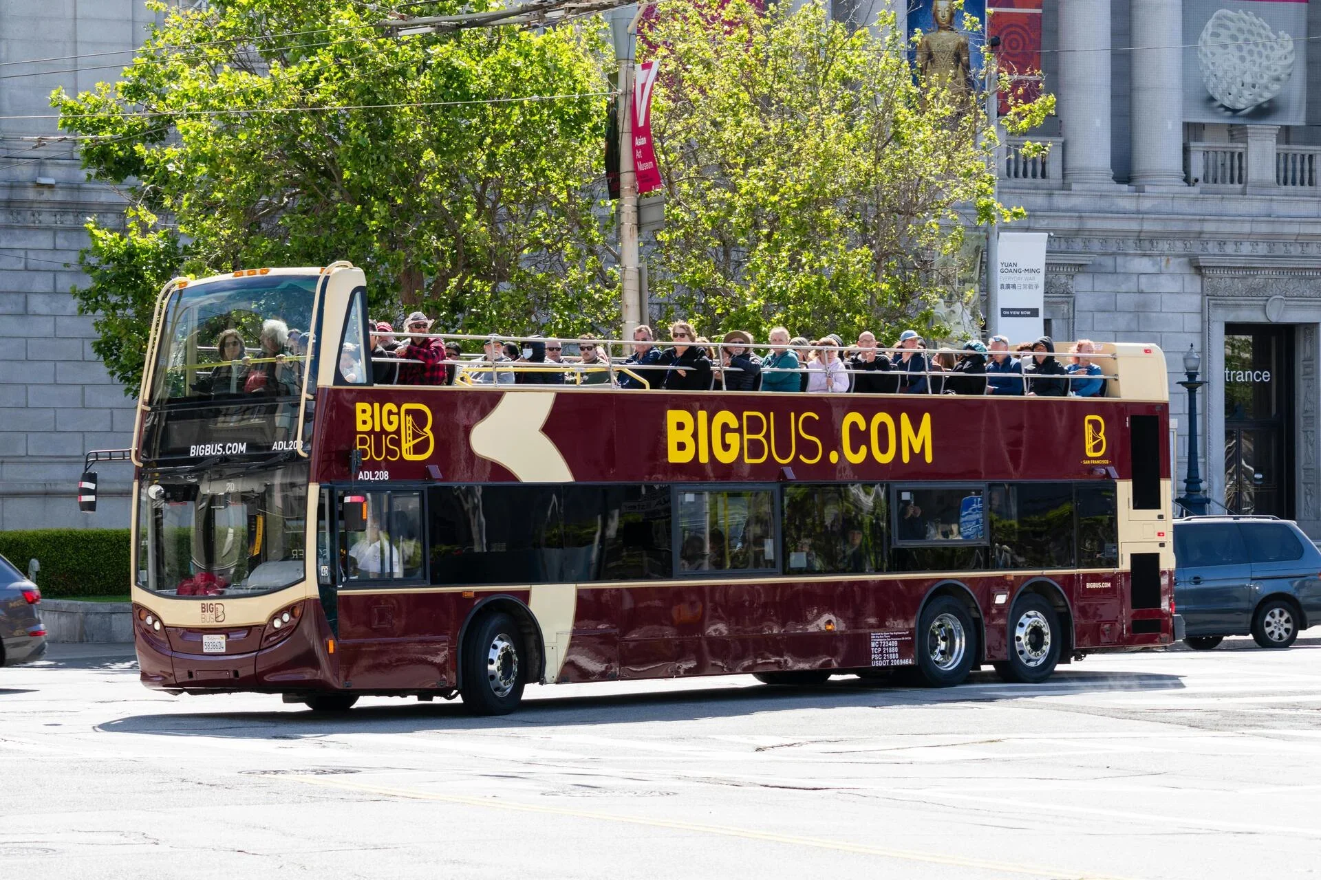 A double-decker bus with open top carrying tourists, driving through a city street with green trees and a historic building in the background.