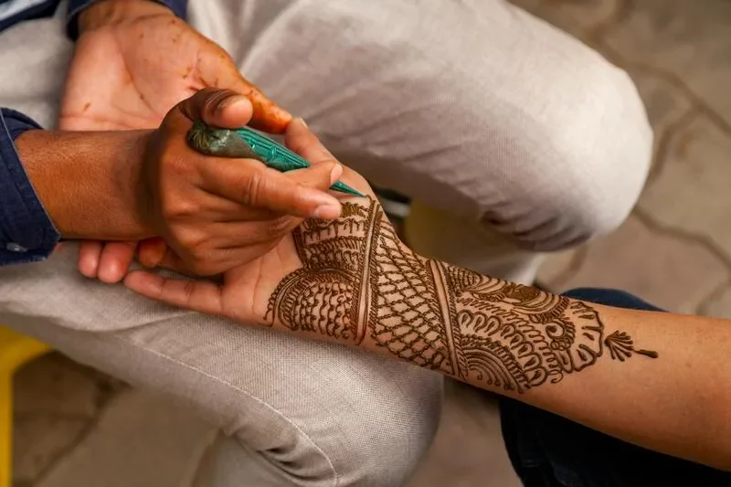 Close-up of a person's hand with intricate henna designs, holding a green tattoo stamp. The person is resting their arm on another person's lap, who is wearing beige pants.