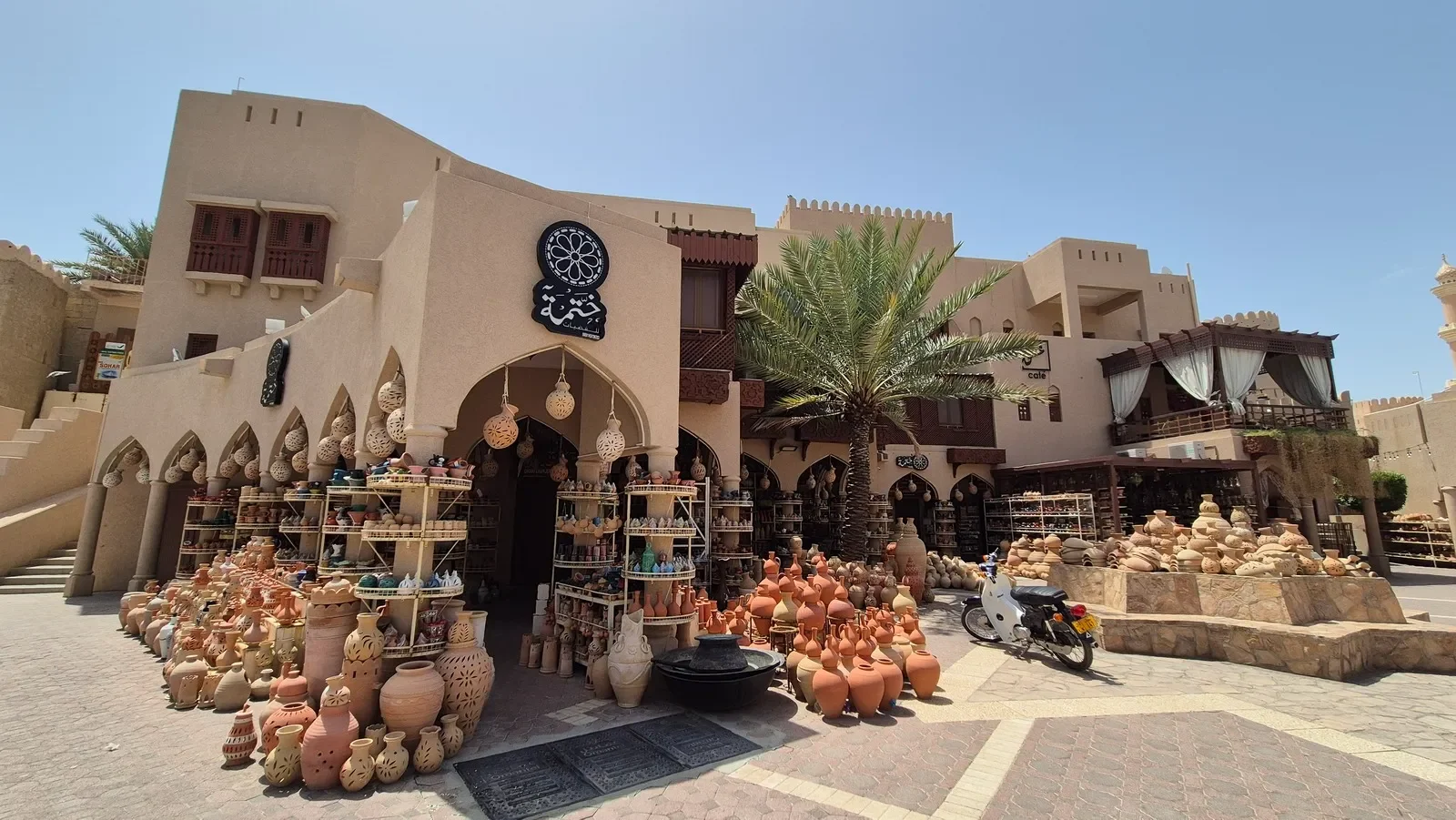 Clay pottery shop with shelves of various vases and pots outside, a palm tree, and a beige building with arched windows and balconies, in a sunny setting.