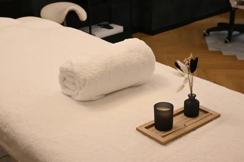 A neatly rolled white towel on a spa bed, with a small decorative arrangement of black and white feathers in a black vase, candles, and a wooden tray with a matchstick on it, in a relaxation or spa setting.