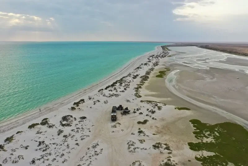 Aerial view of a beach with turquoise water on the left, white sand, and some greenery and tents on the right side.